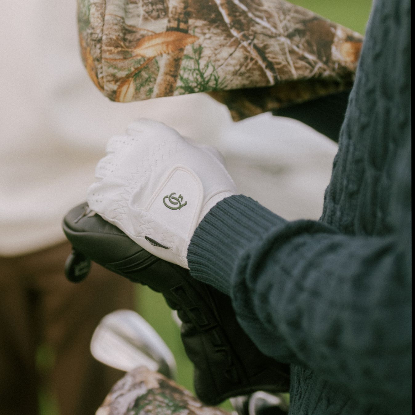 Close-up of a golfer in a forest green knit, holding a golf bag with his white gloved hand.