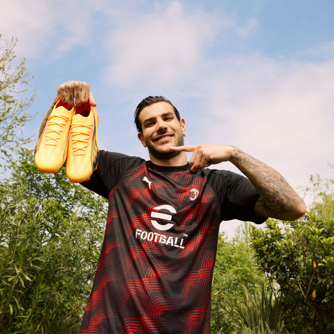 Theo Hernández standing in the park in his football shirt and pointing to his yellow PUMA trainers, which he holds up in the air.