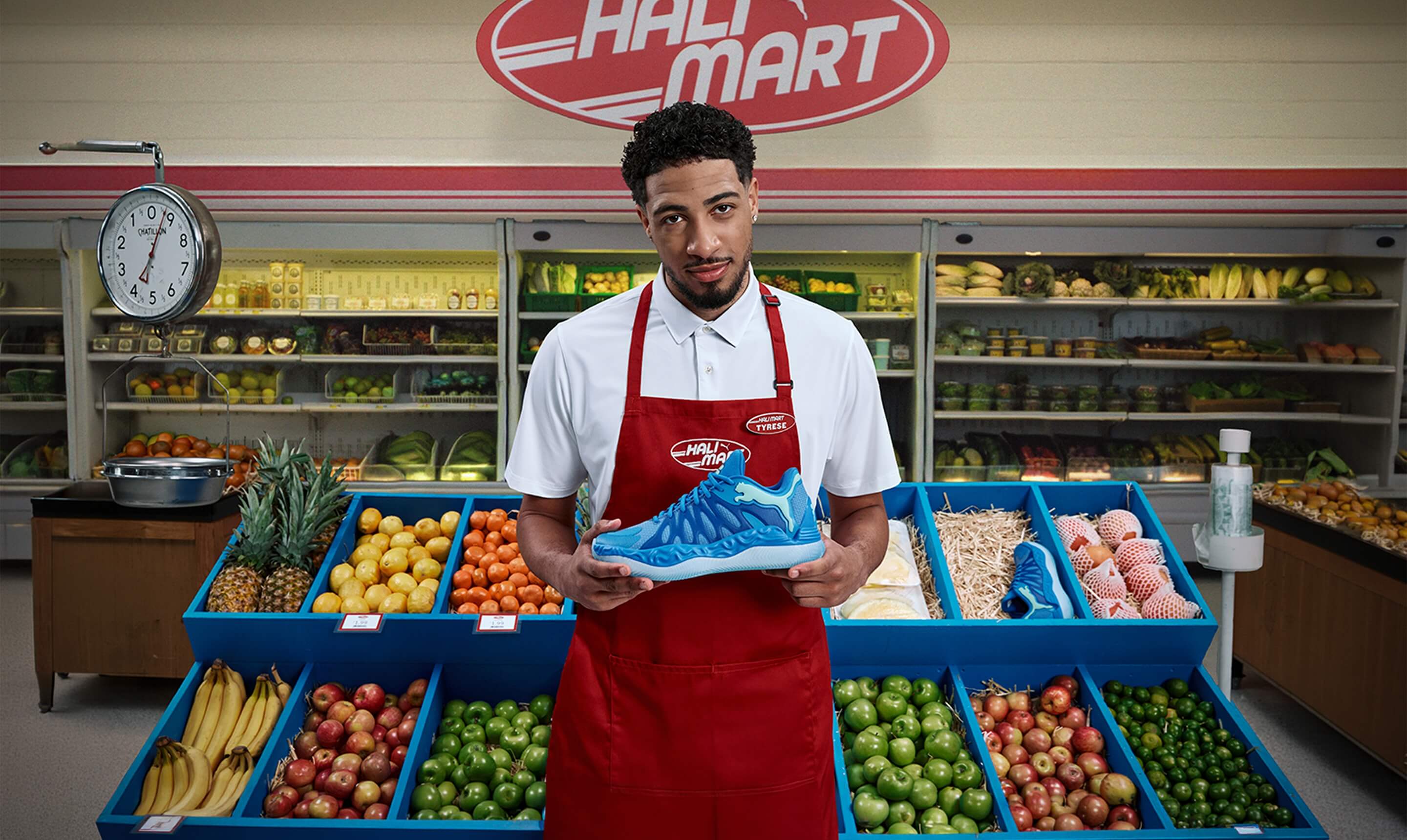 NBA‑Spieler Tyrese Haliburton tr&auml;gt ein Verk&auml;ufer‑Outfit mit roter Sch&uuml;rze und pr&auml;sentiert den blauen Puma Hali 1 Basketballschuh vor einer Gem&uuml;seauslage in einem Supermarkt mit Halimart‑Logo