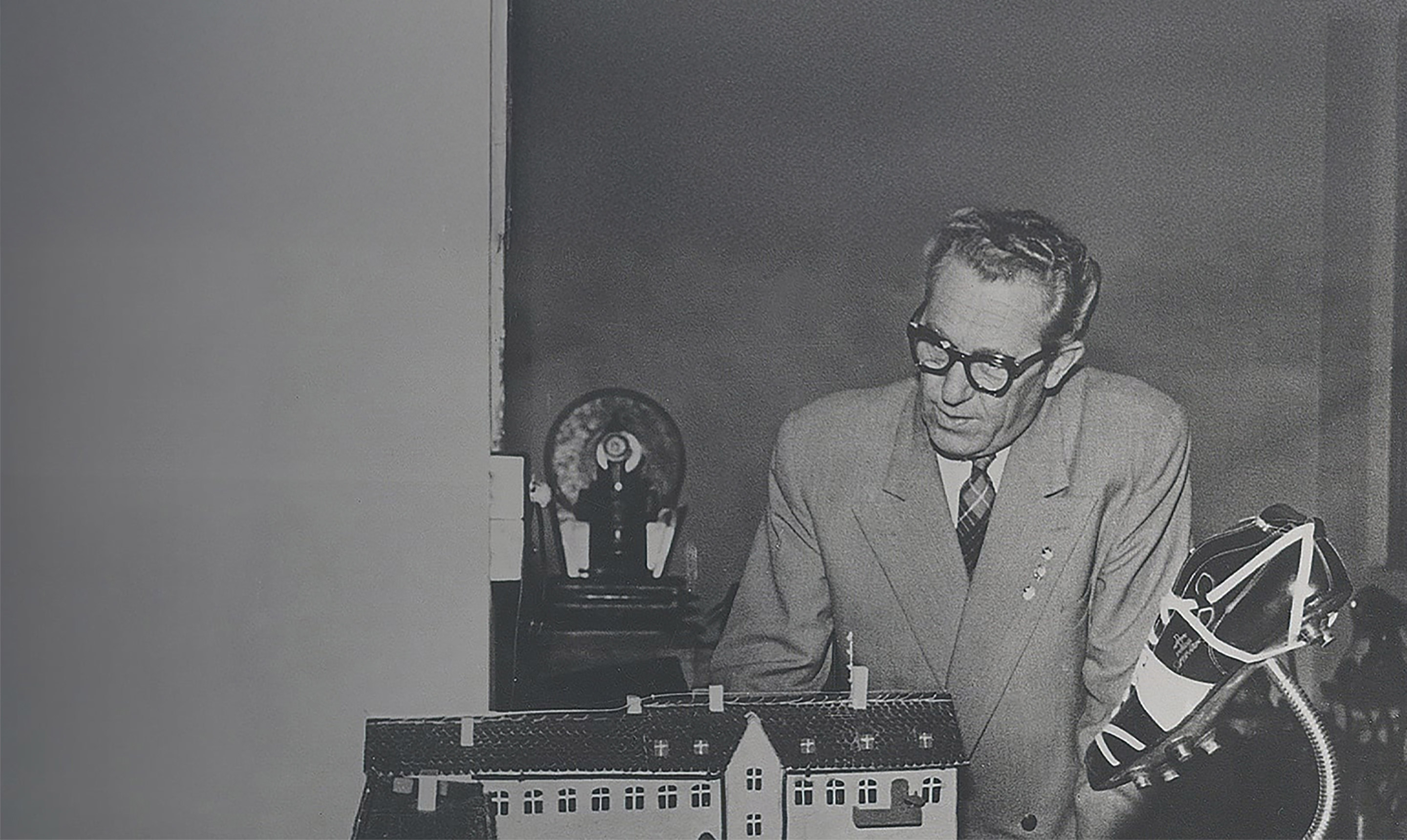 Black-and-white photo shows Rudolf Dassler in a suit beside a detailed building model and an early sports shoe displayed on a stand