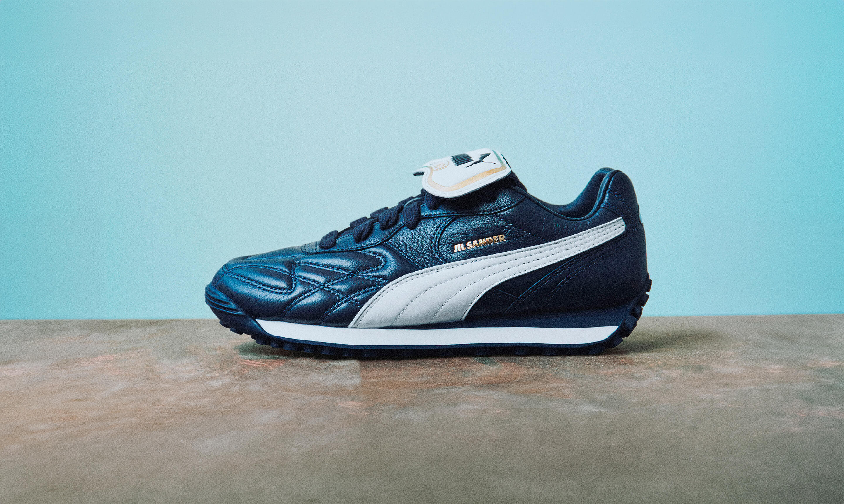Jil Sander x PUMA King Avanti sneaker in dark blue with white formstrip and gold accents displayed side-on against a light backdrop